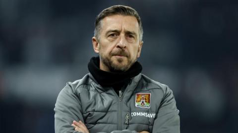 Jon Brady in a grey coat and black scarf on the touchline at a game.