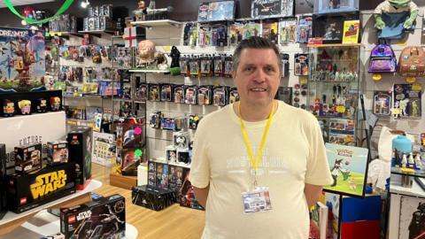 A middle-aged man wearing a pale yellow top which says "Future Nostalgia Toys" and a yellow lanyard smiles as he stands in his toy shop. He has short brown but greying hair. Around him, on display, there are Star Wars Lego sets, Pokemon, and collectible figures.