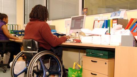 Man with dreadlocks sits at an office desk, in a blue wheelchair, typing at a desktop computer