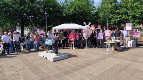A sunny outdoor rally of people holding pink protest signs under a white canopy tent in a paved area. A pink mascot stands in the crowd, a small statue wrapped in black plastic sits in the foreground, and green trees line the background.
