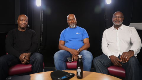 Three people sit on chairs facing the camera in a studio setting, with lights behind them and a small table in the foreground.