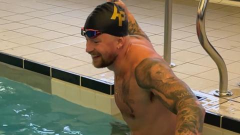 Adam Peaty smiles as he climbs into a pool wearing a black swimming cap with AP in gold letters and swimming goggles on his forehead.