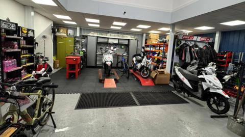 A picture of the warehouse. It has a number of bikes with repair stations. There is items around the warehouse, with motorbikes also in the distance.