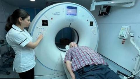 An older man wearing a purple and grey checkered shirt goes in for a CT scan as a nurse watches over him