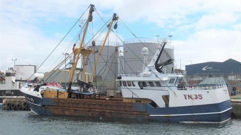 Scallop-trawler Olivia Jean in a harbour, with blue hull and yellow rigging, with industrial buildings in the background