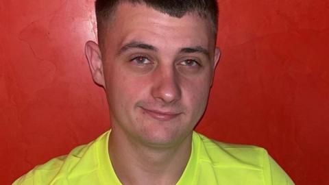 Fraser Stewart is half smiling at the camera. He has short, brown hair, shaved at the sides and is wearing a luminous yellow T-shirt. He is standing against a red wall.