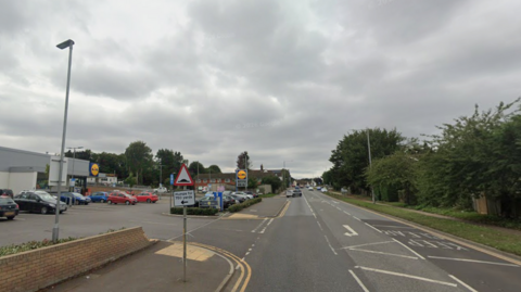 A road with a Lidl supermarket on one side and a row of fences and trees at the other.
