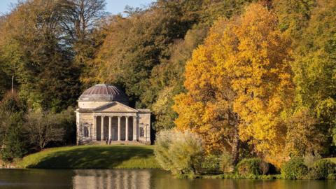 Stourhead in Wiltshire. There are orange, green and yellow trees in the shot and a building overlooking water. 