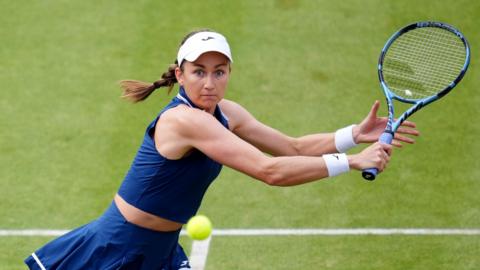 Olivia Nicholls playing at Queen's. She is dressed in a navy sports outfit and is wearing a white cap. She is preparing to play a backhand shot and is looking intently at the yellow ball.