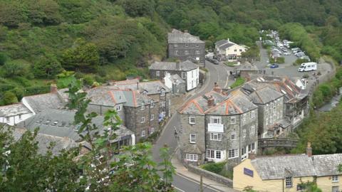 The village of Boscastle is seen from higher up - there are cars parked at the top of the village with a road sweeping down between the house and the river to the right of the houses