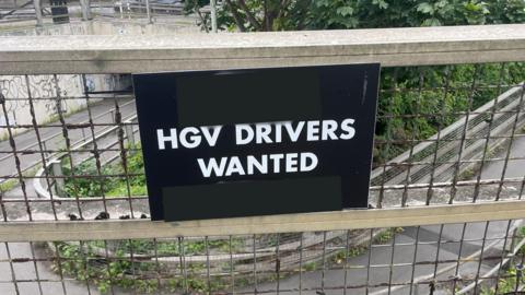 A black sign reading "HGV drivers wanted" in white capital letters. It is stuck to a  metal fence.