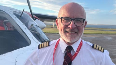 A smiling male pilot, who is bald with a short white beard and is wearing a white shirt and a red tartan tie, stands next to a small aircraft. It is a bright sunny day and the coast is visible in the background, beyond the tarmac.
