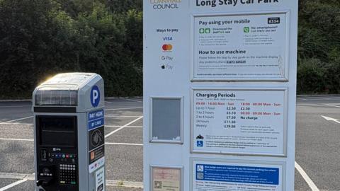 A payment machine on the left next to a white board explaining the various payment methods and tariffs in a Cornwall Council car park. 