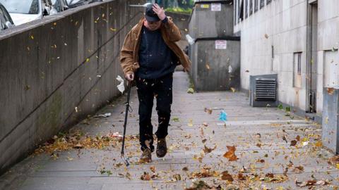 Man walking with a stick on a street being buffeted by strong winds with leaves swirling around his feet
