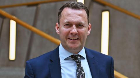 Russell Findlay, who is wearing a midnight blue suit jacket, a light blue shirt and a black tie, looks into the camera smiling