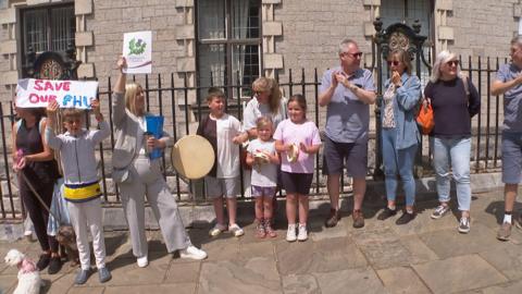 Protestors gather in Torquay ahead of council decision. There are both male and female protestors present. They are banging instruments and holding up banners. Young children are in attendance.