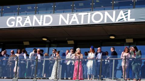 Smartly-dressed spectators look out from a balcony under a large banner saying "Grand National"