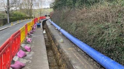 A road in the Bradford area with security barriers at one side with a long hole being prepared to receive a new water pipe