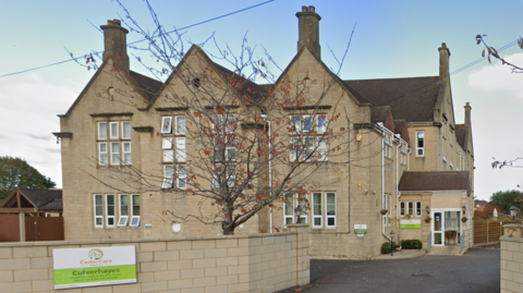 External shot of Culverhayes Nursing Home, which is a sand-coloured building. A wall with a sign saying ' Culverhayes' is on a surrounding wall.