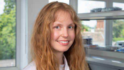 Dr Lindell is in a lab with windows behind her. She is smiling at the camera while wearing a white lab coat. She has long ginger hair and light blue/ green eyes. 