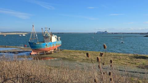 A small fishing trawler sits on a shoreline before a calm sea, under a blue sky
