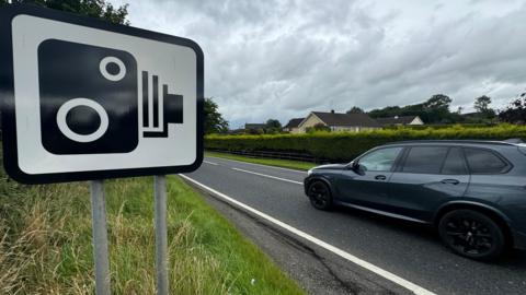 A black and white speed camera road sign on a grass verge at the side of a road, with a residential area in the background. A dark grey vehicle is travelling past the sign.