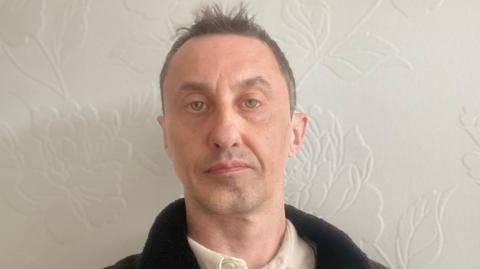 David Robinson is photographed indoors against a pale wall with a subtle floral pattern. He is shown from the shoulders up, wearing a light shirt and a dark jacket. The lighting is soft and even, and the background is uncluttered.