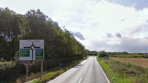 Long straight country road with a road sign on the left showing roundabout graphic with town names