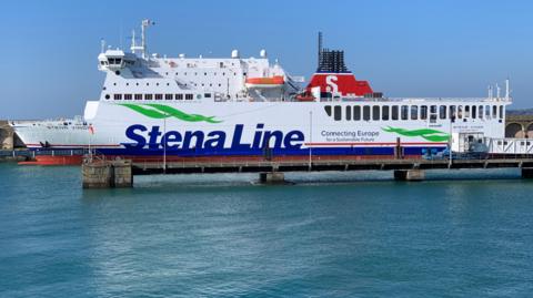 The Stena Vinga ferry is coming in to port. It has Stena Line Connecting Europe for a Sustainable Future printed on its port side. The sky is blue and the sea is calm.