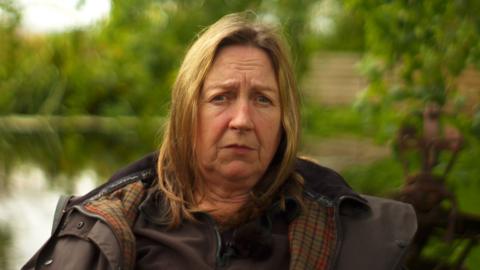 A woman with dyed blonde hair and blue eyes sits in front of a blurred outdoors background with lots of trees and bushes. She wears a brown coat with red and green tartan lining and a brown fleece underneath.