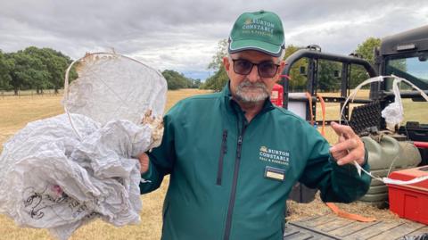 Wildlife officer Dave Nassau. He is wearing a green jacket and cap and is holding the remains of two sky lanterns. There is an expanse of brown-coloured grassland behind him.