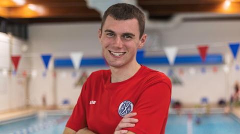 Luke Brown smiles as he looks directly into the camera. He is standing next to a pool at Penrith Leisure Centre. He has short dark hair and is wearing a red T-shirt bearing the logo of Penrith Swimming Club.