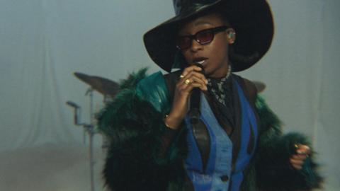 Little Simz performing at the BBC's Maida Vale studios. She wears an oversized leather top hat, blue leather waist coat, green furry coat and dark sunglasses. She raps into a microphone while looking over her right shoulder against a white backdrop. 