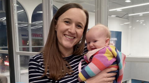 Dark haired woman wearing a stripey black and white top smiling, holding a small baby in a stripey multicolored knit top