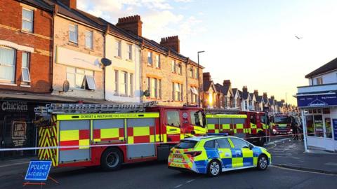 Several fire engines and a police car blocking a road of shops with flats above.