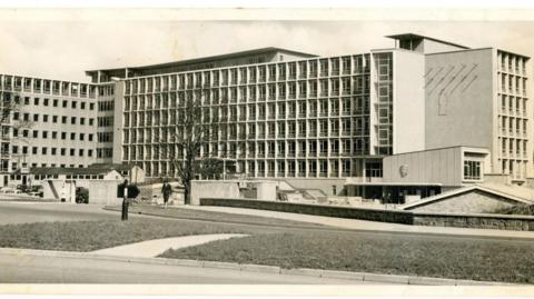 A black and white photograph of Durham County Hall. The tall beige building is boxy, covered in windows and surrounded by a grassed area.