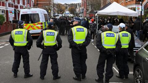 Five police officers stand in a line in a street with a police van in front of them as a crowd of people, some wearing COVID face masks, gather in front of them.