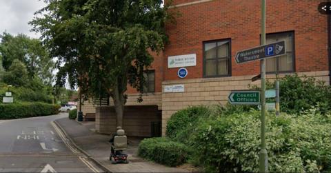 A red-brick Three Rivers District Council building stands beside a tree-lined road. Signs attached to a lamppost direct visitors to Watersmeet Theatre and the council offices. A person on a mobility scooter travels along the pavement in the foreground, with hedges and mature trees framing the scene.