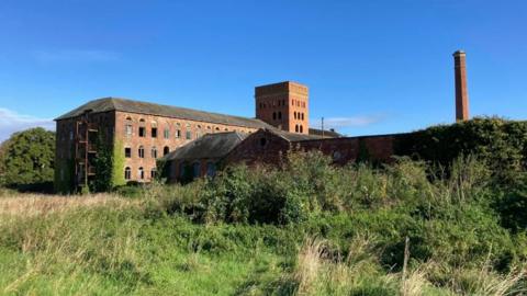 An exterior shot of the Tone Works complex in Wellington, Somerset. The 18th century factory buildings are in a visible state of disrepair. Windows are broken and shrubbery is climbing a building in foreground. Over grown grass and climbing Ivy paints the site as entirely derelict.   