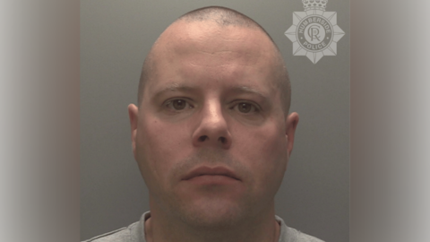 A police custody shot of Ian Walpole. He has a bald head and is looking at the camera. He is wearing a light grey t-shirt. The wall behind him is grey.