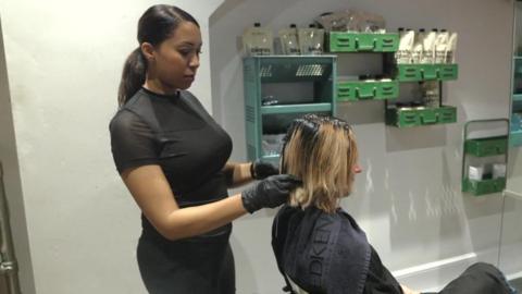A young woman in a hairdressing salon wearing gloves putting colour in the hair of a blond woman sat in a chair