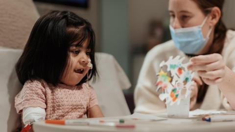 Zarwa is pictured sitting at a hospital table smiling as a masked staff member helps her make a colourful paper snowflake during an arts and crafts activity.