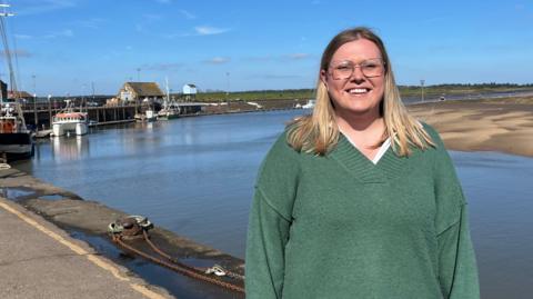 Annie Golding stands on Wells quayside in the sunshine. She has shoulder length blonde hair and is wearing a green jumper. Behind her is the sea and sand dunes. 