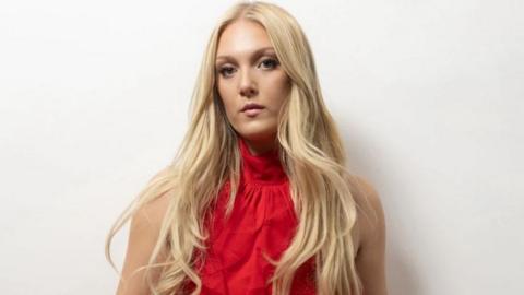 Emily Fern, a woman with blonde hair who is wearing a red top. She is looking at the camera with a neutral face.