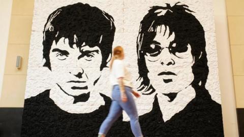 A woman walks past a large black and white mural of Noel and Liam Gallagher made out of bucket hats. 
