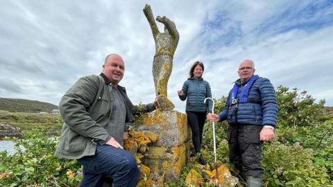 Dithis fhear agus boireannach nan seasamh ri taobh na h-ìomhaigh concrait a th' ann