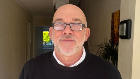 A man with balding grey hair and a beard and wearing a white shirt and dark coloured jumper is smiling as he stands inside the hallway of a house.