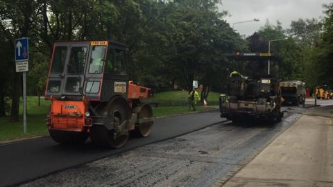 A roller vehicle parked on some newly-laid tarmac on a road shut off for resurfacing. Another work vehicle can be seen close by as workmen walk around the site, which is surrounded by trees.