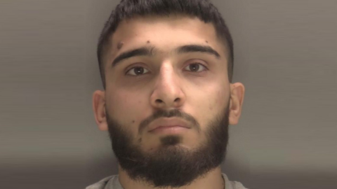 A close-up of a man's face. He has a moustache and beard and is looking straight at the camera, and is pictured against a grey background