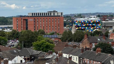 A large building can be seen rising up amongst rows of terraced houses, sitting next to a multi-coloured glass building